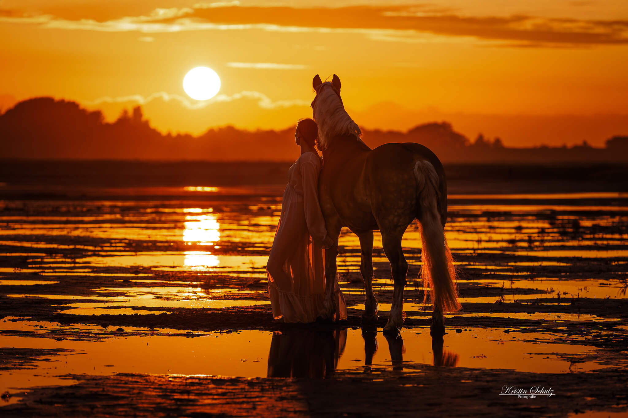 Eine Frau steht mit einem Pferd in der Dämmerung, der orangefarbene Sonnenuntergang erleuchtet die Szene.