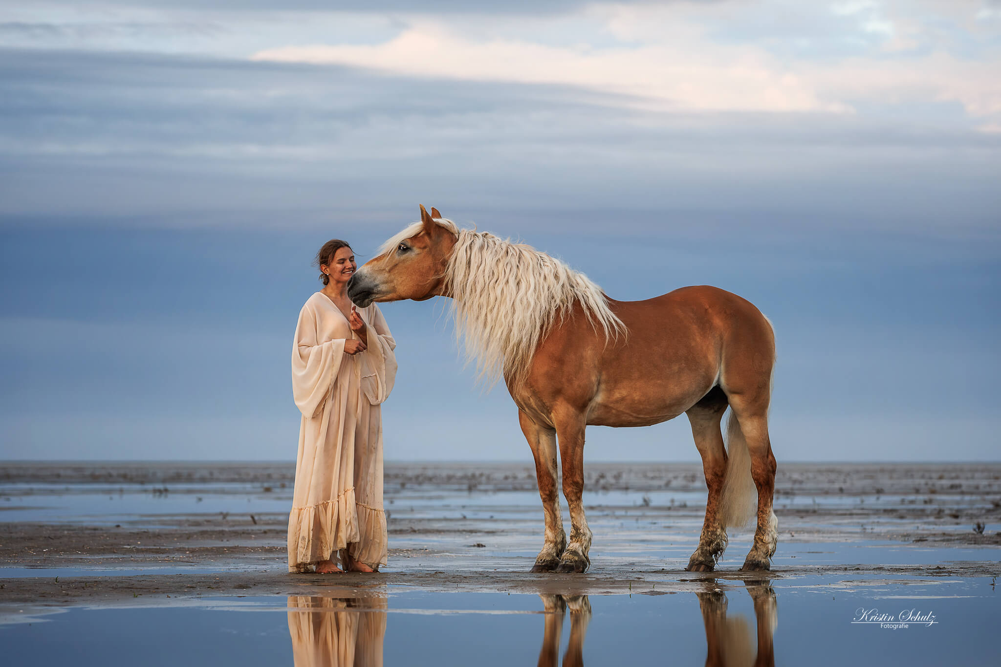 Verbindung zwischen Mensch und Pferd im ruhigen Wasser Eine Frau steht mit einem Pferd am Ufer eines Sees, die beiden blicken sich gegenseitig an, während sich der Himmel im Wasser spiegelt.