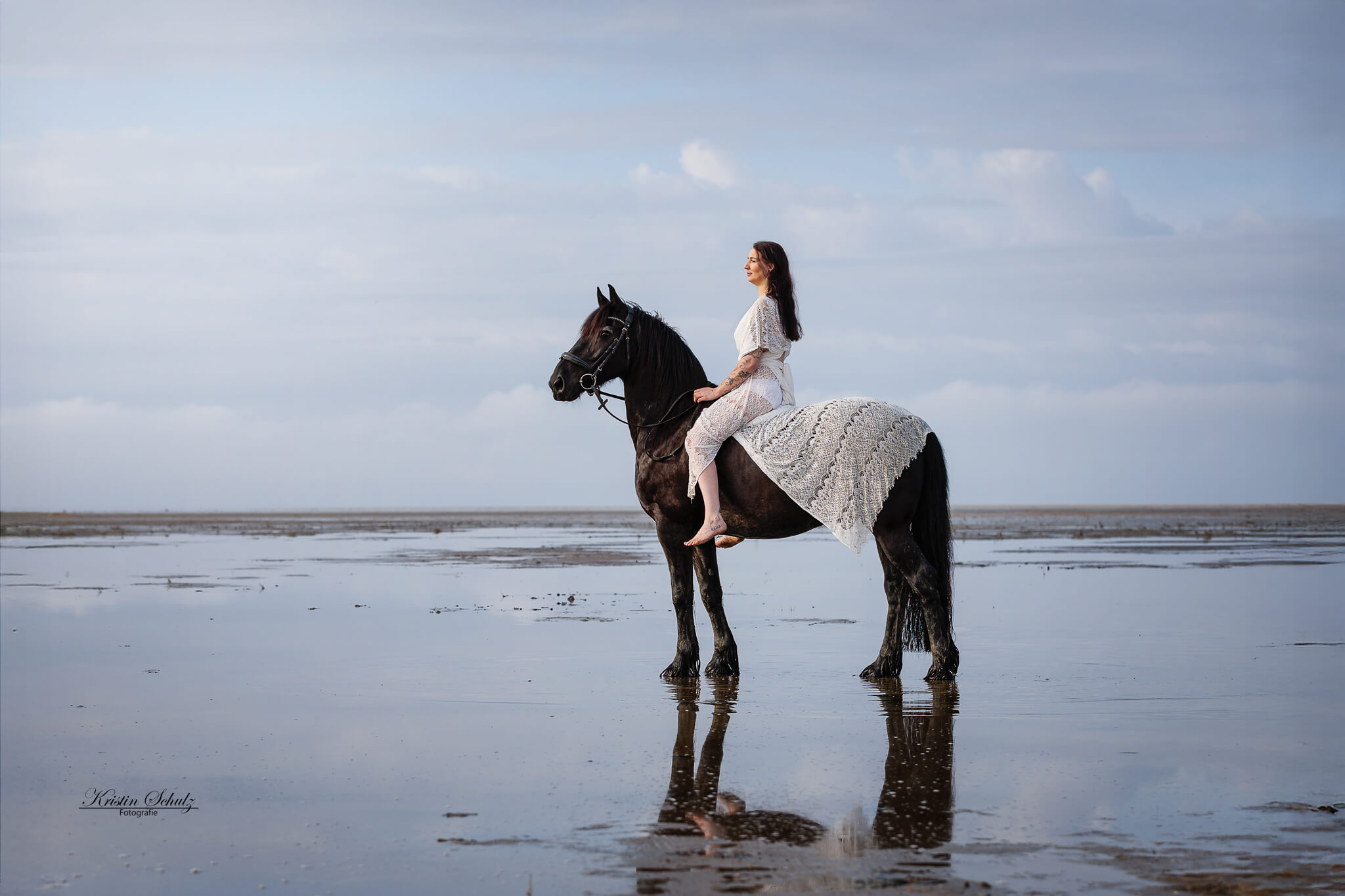 Schwarzes Pferd und seine Reiterin stehen am Strand und schauen der Sonne entgegen