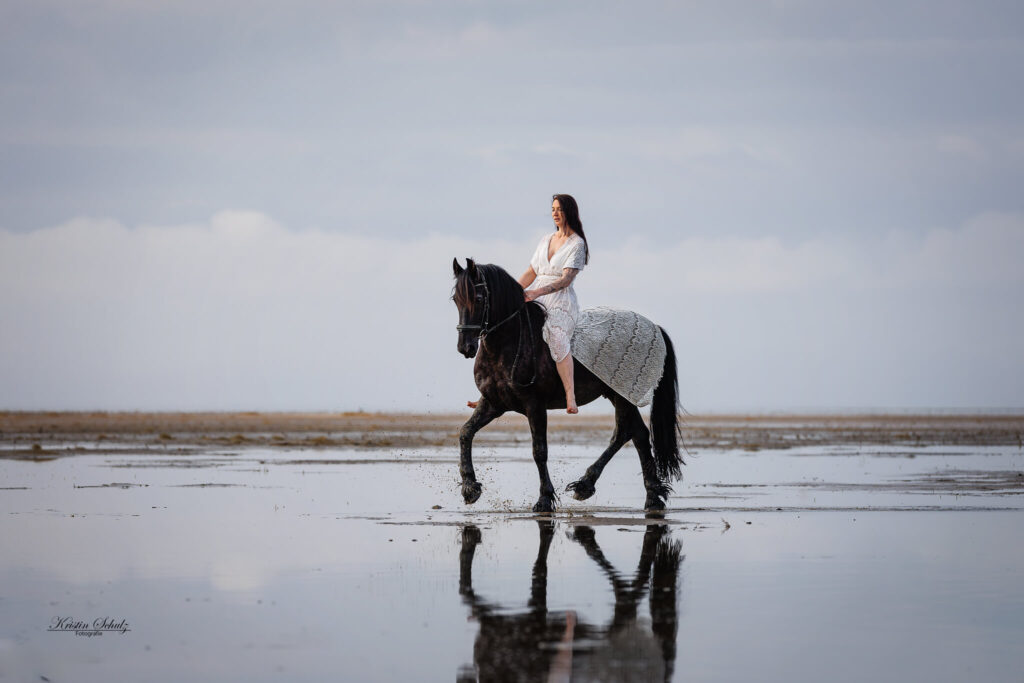 Schwarzes Pferd trabt mit Reiterin durchs flache Wasser am Strand
