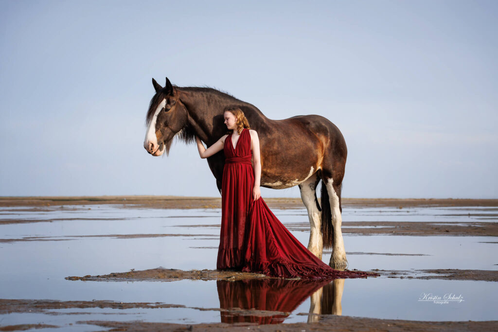 Ein junges Mädchen in einem roten Kleid steht mit ihrem Pferd in einer Wasserfläche, die das Bild spiegelt.